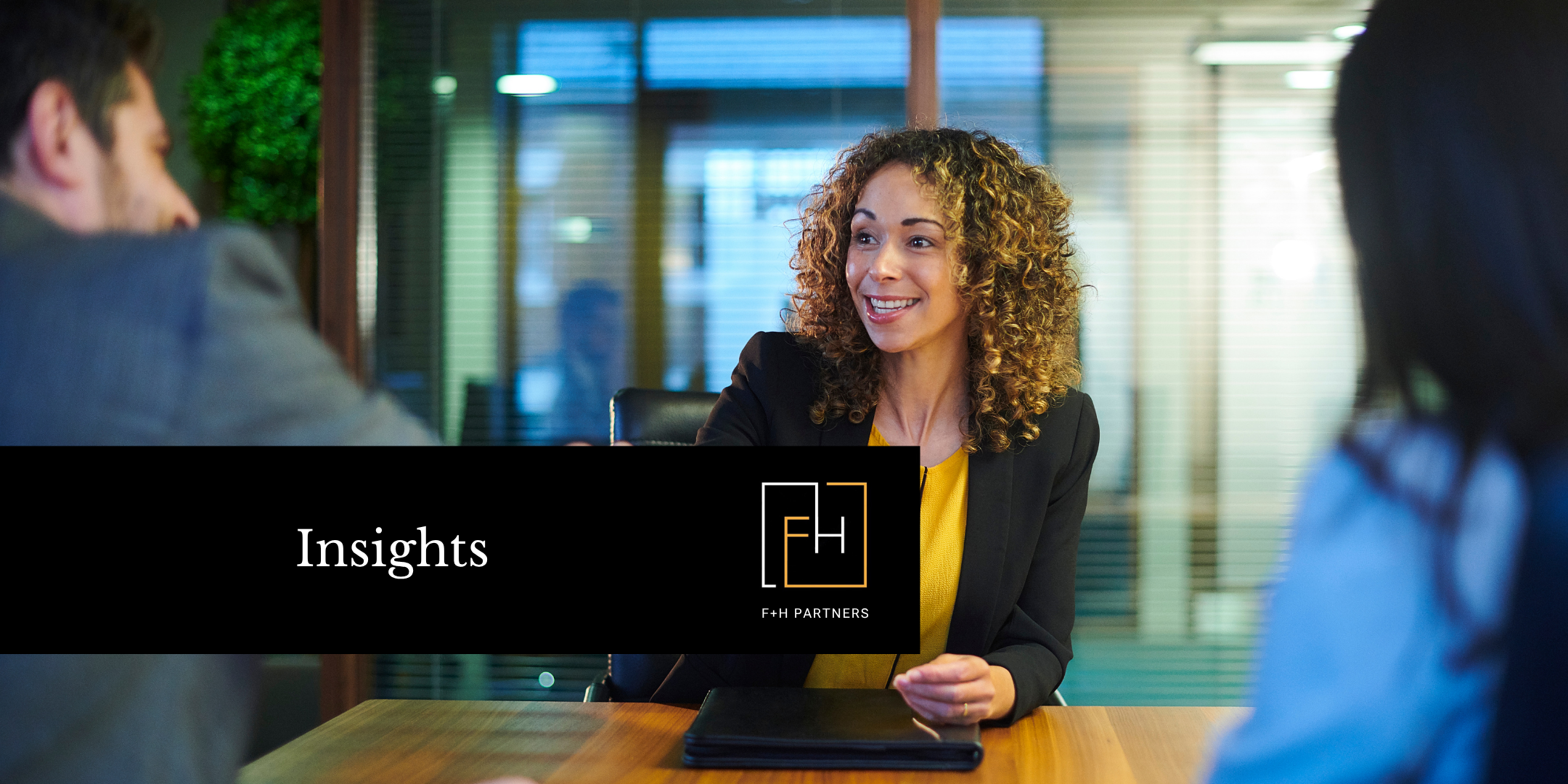 A professional woman smiles and reaches across a table to shake hands during an interview or meeting.